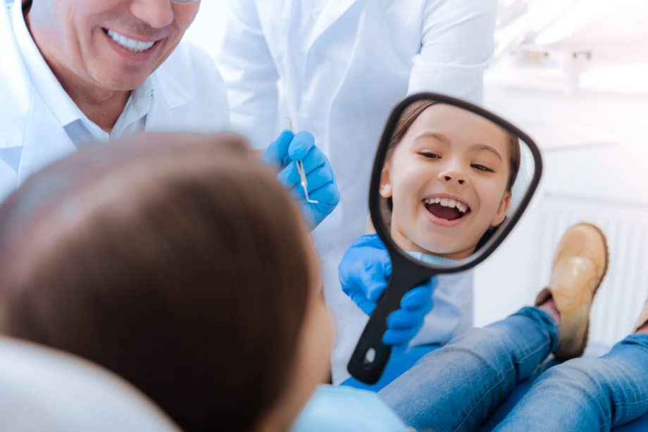 Happy child at the dentist, smiling while looking in a hand held mirror while a male dentist smiles beside her.
